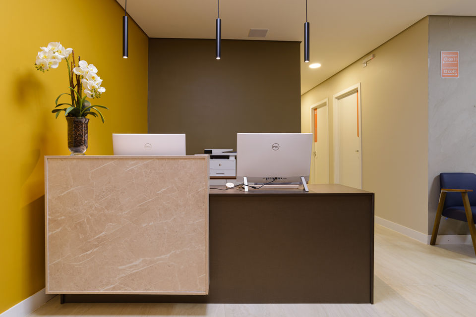Recepção de consultório com balcão em mármore bege e superfície marrom, dois computadores brancos e vaso com orquídeas brancas. Paredes em tons de amarelo e marrom, iluminação com pendentes pretos e corredor lateral com portas brancas.