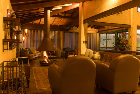 Sala de estar ampla com teto de madeira e paredes em tom bege, decorada com sofás de couro marrom e poltronas confortáveis. Lareira suspensa metálica ao centro, móvel rústico com arranjo de flores tropicais e grandes janelas com cortinas claras que iluminam o ambiente.