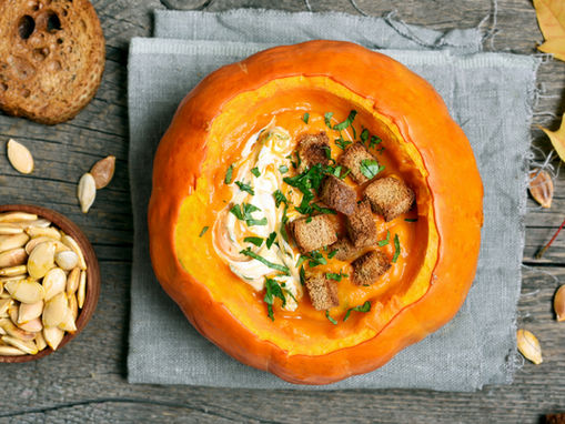 Pumpkin bowl with creamy soup, croutons, and herbs on a rustic wooden table. Accompanied by a bowl of seeds, bread, and autumn leaves.