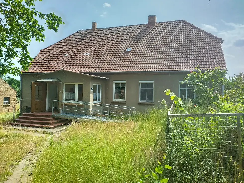Haus mit rotem Ziegeldach, umgeben von hohem Gras und Pflanzen. Die Holztür ist geöffnet, sonnige, ruhige Atmosphäre. Keine Menschen sichtbar.
