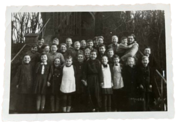 Eine Gruppe von Kindern und einem Erwachsenen posiert draußen für ein Foto. Sie lächeln, die Stimmung ist fröhlich, Bäume im Hintergrund. Eine Dorfschulklasse aus fer 1940er Jahren.