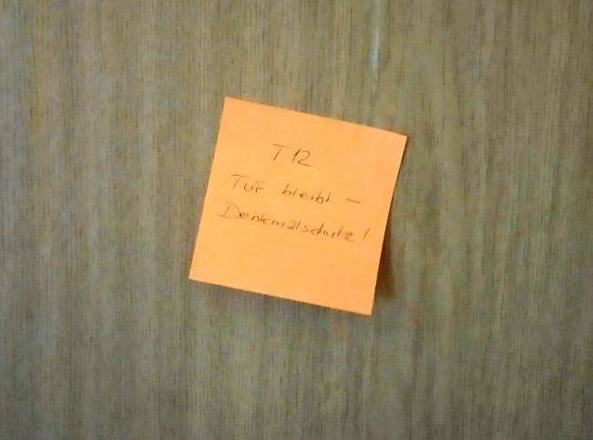 Oranger Klebezettel auf Holzfurnier mit Aufschrift "T12 Tür bleibt - Denkmalschutz!"