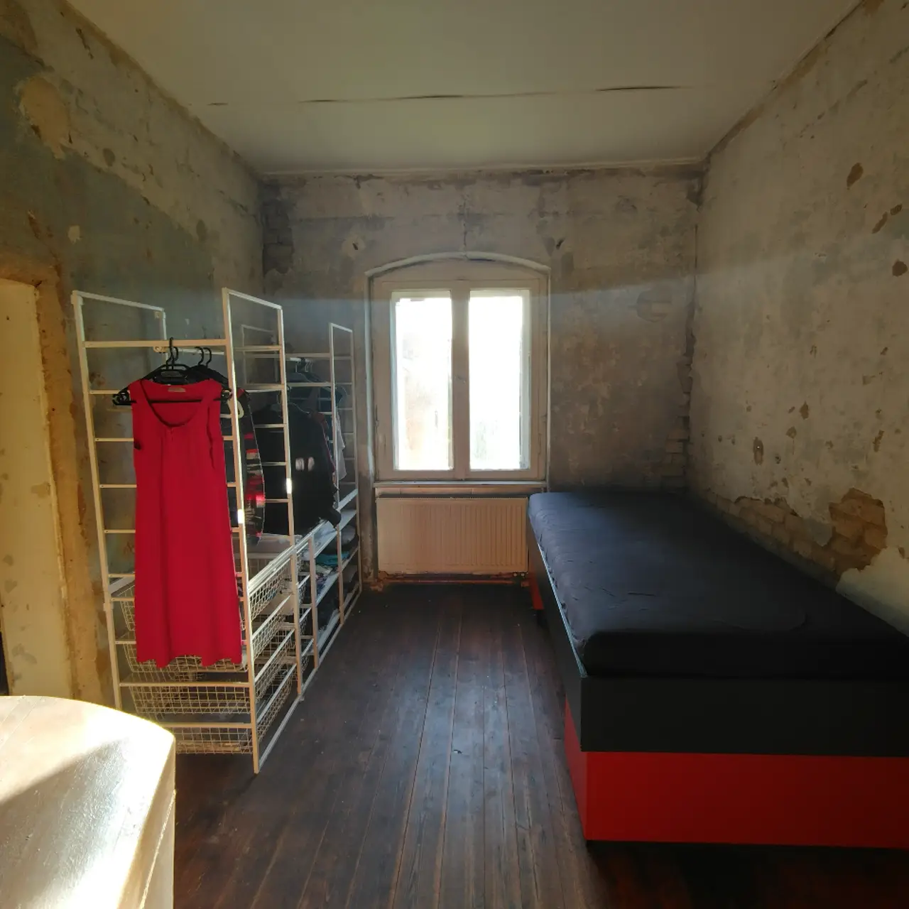 Halbrenoviertes Zimmer mit abgeschlagenem Putz an den Wänden, Kleiderstange und Einzelbett