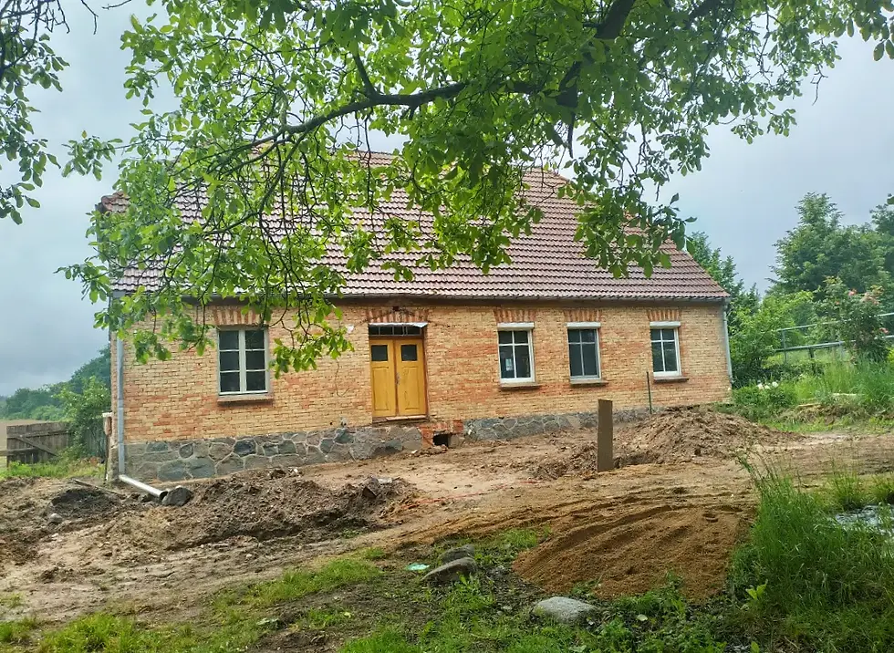 Backsteinhaus mit braunem Dach und gelber Tür in grüner, ländlicher Umgebung. Baumzweige verdecken teilweise die Ansicht. Baustelle davor.