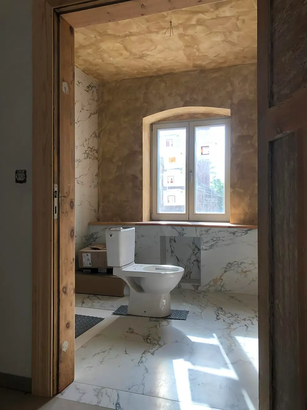 Neues Badezimmer mit Marmorboden und Toilettenschüssel, Fenster mit Sicht nach draußen. Licht fällt durch das Fenster.