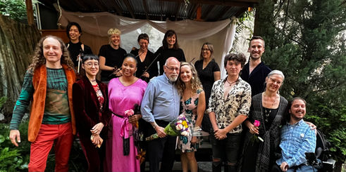 A gathering of brightly dressed people stand together before a wooden open air stage as a cast, crew and creators of an open air opera complete a successful 4 show run. 