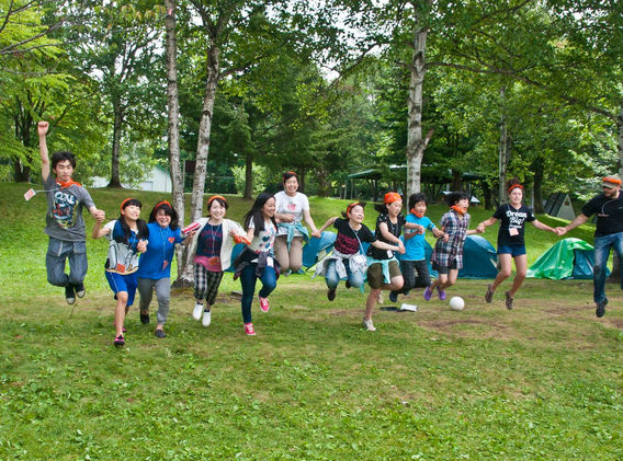 A group of kids jump in the air holding hands on a grassy hill in front of some camping tents.