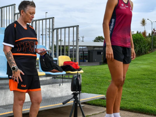 An athletic woman in a maroon Queensland sports singlet and black shorts stands on a wooden box wearing white socks, preparing for a fitness test. A coach beside her, dressed in a black and orange TFC uniform, holds a device and observes. A camera or sensor on a tripod is positioned in front of them, with benches and grassy sports grounds in the background.