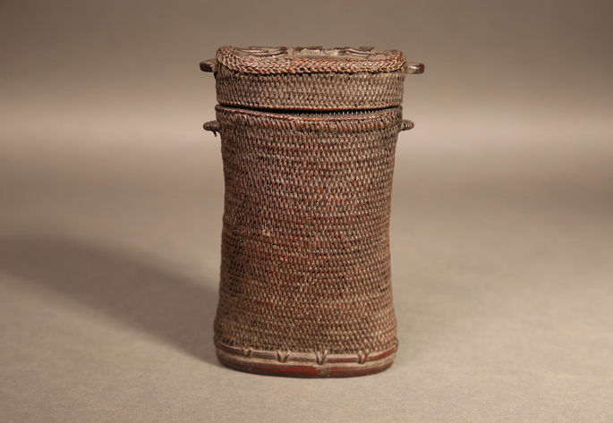 A finely woven, small, cylindrical rattan basket with a carved anthropomorphic figure on its lid