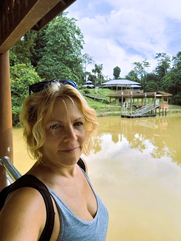 Egy nő kék trikóban mosolyog egy tó partján, háttérben zöld fák, épületek és lépcső. Derűs, nyugodt vidéki hangulat.