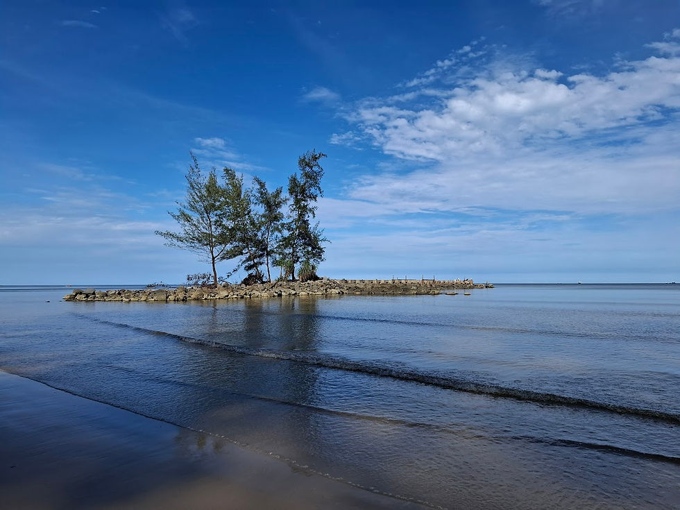 Tiszta kék ég alatt, tengerparti sziklás fövenyen fák állnak. Nyugodt vízfelszín, derűs hangulat. Felhős égbolt a háttérben.