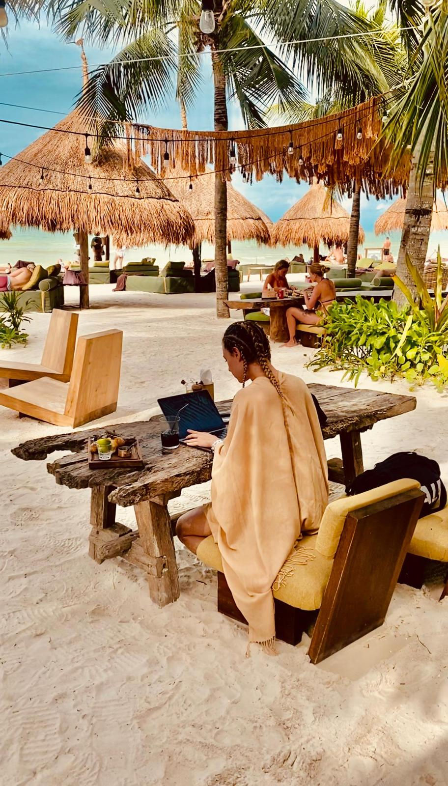 Life coach working remotely on a laptop at a tropical beach resort, showcasing a lifestyle of abundance and freedom.