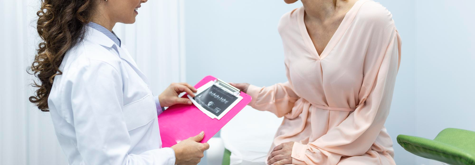 Médica mostra ultrassom a gestante sorridente em consultório claro, com prancheta rosa e maca verde.