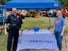 Kat Challis and Carroll Berg from NH Children’s Trust attend National Night Out events in Concord and Goffstown. Kat staffs a booth at Rollins Park with family resources and colorful materials. Carroll stands behind an outreach table at Goffstown High School, engaging with community members. Families and children explore activities, trucks, and community exhibits in the background. The events highlight NH Children’s Trust’s work to support strong families and resilient communities.