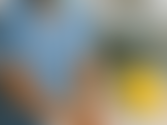 Healthcare worker in blue scrubs and a surgical mask washing their hands at a sink, with a small yellow sharps bin visible on the counter, in a blurred hospital treatment room background.