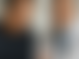 A concerned middle-aged man in a dark sweater listens intently as a doctor in a white coat and stethoscope explains medical results. The doctor holds a document and gestures empathetically during their conversation in a bright, clinical setting.