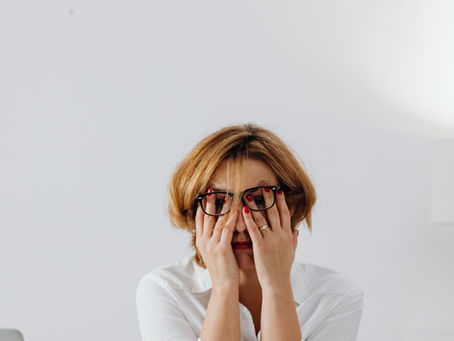 Eine erschöpfte Frau die bei ihrem Schreibtisch vor dem Laptop sitzt und sich die Hände vor das Gesicht hält.