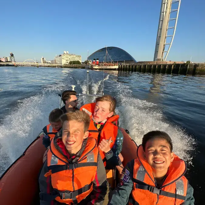 Children smiling while on a boat trip on the Clyde. In the background we can see the Glasgow Science Centre, BBC Studios, and the Clyde Arc Bridge.