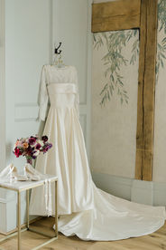 A wedding dress hung by a mirror with bouquet and white shoes