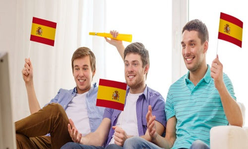 Three men cheerfully wave Spanish flags while watching TV. One holds a yellow horn. They sit on a couch in a bright living room.