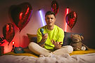 Kevin Neon sitzt auf einem Bett mit einem alten Telefonhörer in der Hand, umgeben von roten Herzballons, einem Teddybär und neonfarbenen Lichtern, in einem stimmungsvoll beleuchteten Raum.