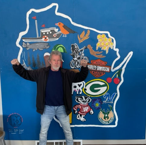 A man standing in front of a blue wall with Wisconsin painted on it with his arms in the air.