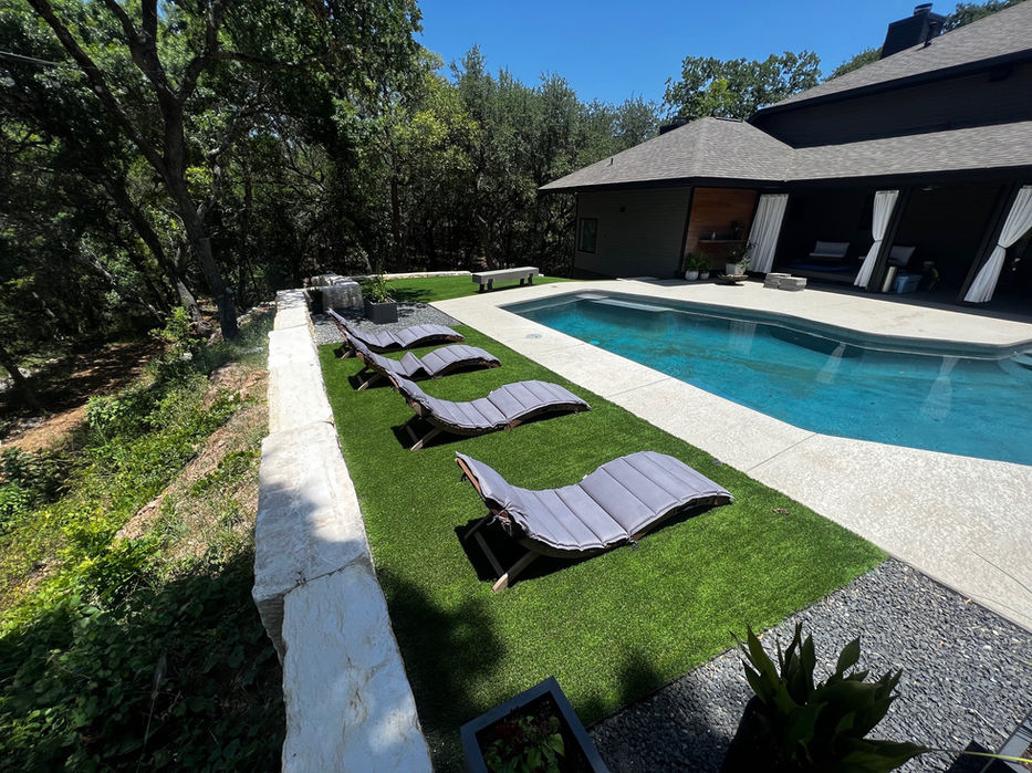 Modern house with deep, open patios.  Lovely, clear pool with turf surrounding.  Small rock bed.  Tall trees and bright blue skies.