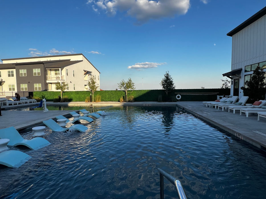 Large pool with several lawn chairs and apartment building in the back.  Artificial Ivy Privacy Wall along side of pool.  Blue skies and white clouds.