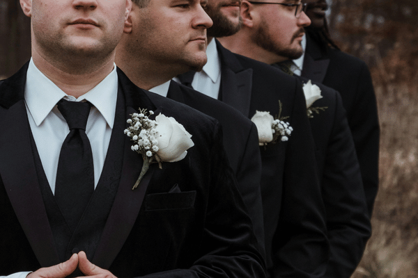 Row of men in black suits with white boutonnieres stand solemnly outdoors against a blurred brown background.