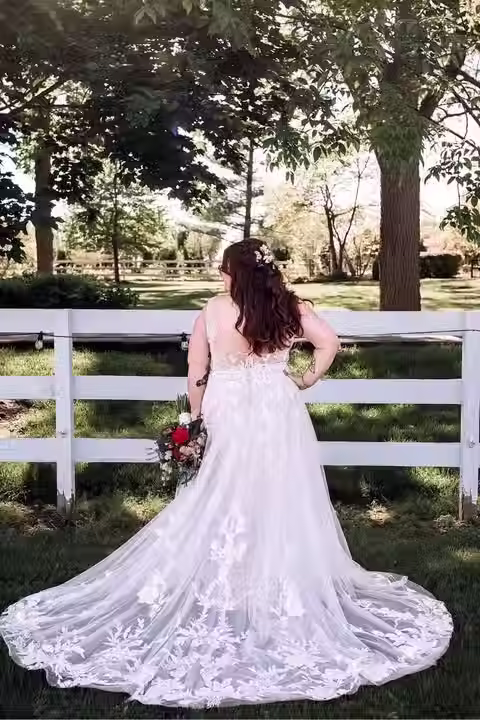 Real Bride Emma stands outdoors facing a white fence, wearing a floral lace A-line wedding gown with a long train and holding a bouquet in a sunlit garden setting.