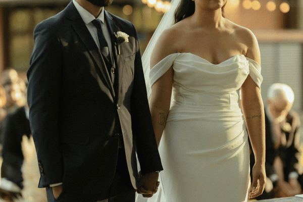 Bride in white off-shoulder gown and groom in dark suit hold hands at a warm-lit wedding ceremony