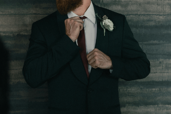 Bearded man in a black suit adjusts his tie, with a white boutonniere, against a dark textured backdrop.