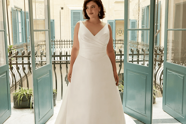 Bride in a white ball gown stands in a sunlit doorway framed by open blue shutters and balcony railings.