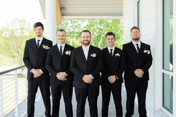 Five men in black suits stand on a porch, hands clasped, with pink and white boutonnieres against a bright green background.