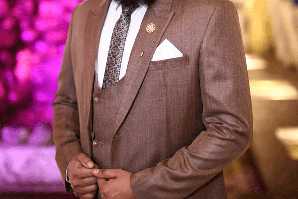Close-up of a man in a brown suit with patterned tie and white pocket square, adjusting his jacket against a pink-lit background.