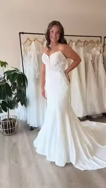 Bride wearing a fitted mermaid wedding dress featuring a lace sweetheart bodice, illusion plunge neckline, and flowing train, posing inside Blush & Ivory Bridal with designer wedding gowns in the background.