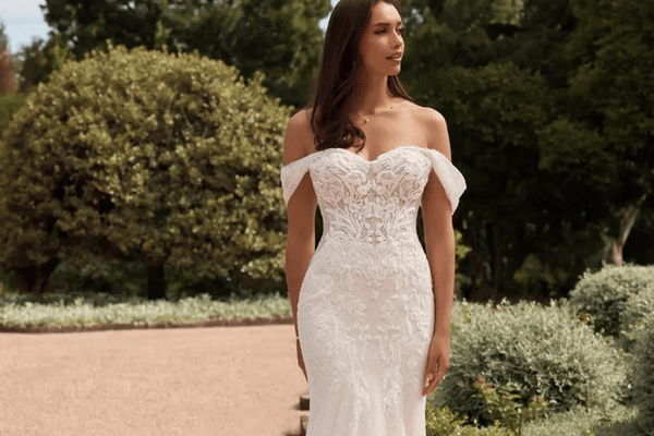 Woman in a lace off-shoulder wedding dress stands in a garden setting. She's looking to the side, exuding a serene and elegant mood.