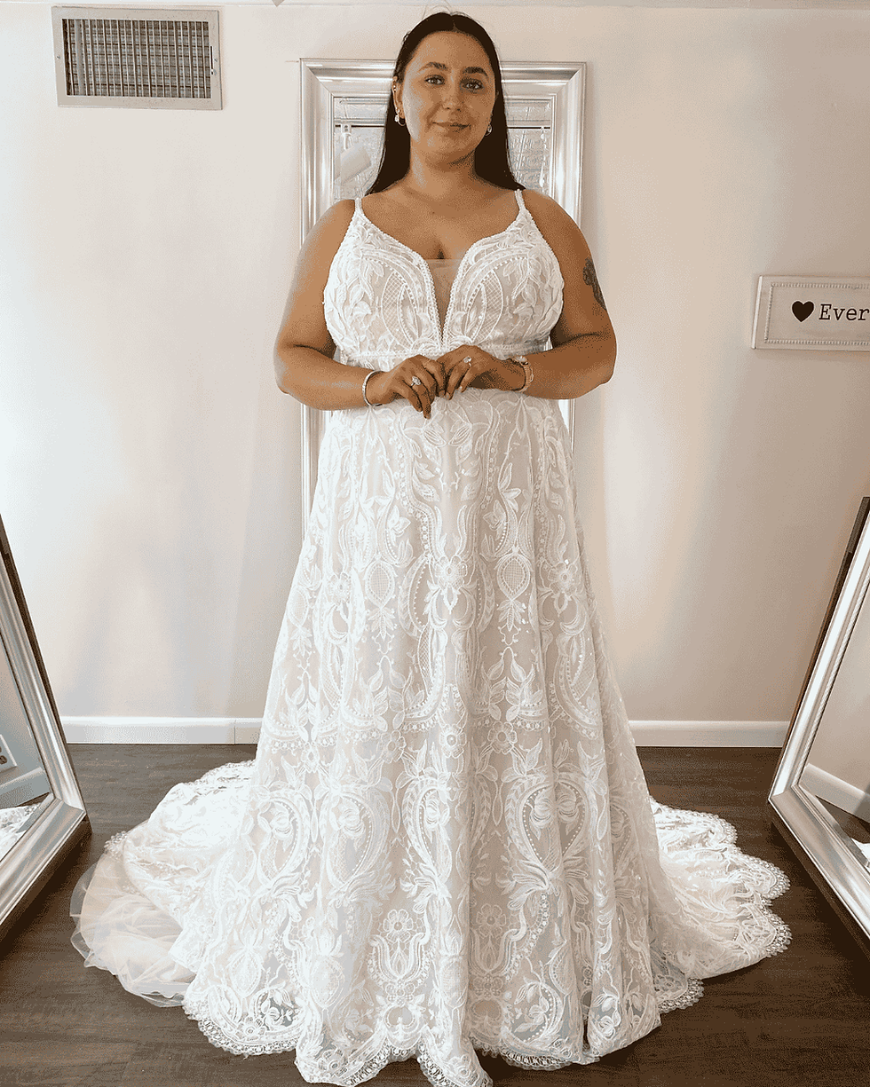 A woman in a white lace wedding dress, styled as an elegant empire gown, stands in a bright room with mirrors.