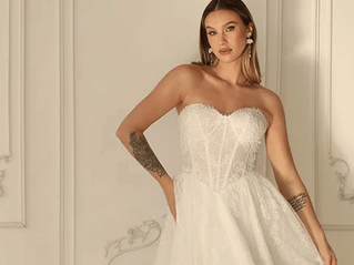 Bride posing in a strapless white lace wedding gown against an elegant cream paneled wall, with a confident, poised expression.
