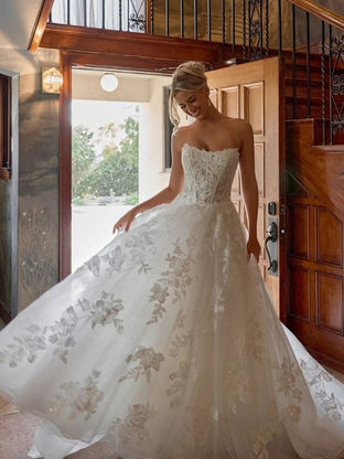 Bride wearing Essense of Australia D4403 strapless leafy lace ballgown wedding dress with a sweetheart neckline and voluminous skirt, featuring floral appliqués and soft tulle layers in a sunlit doorway setting.