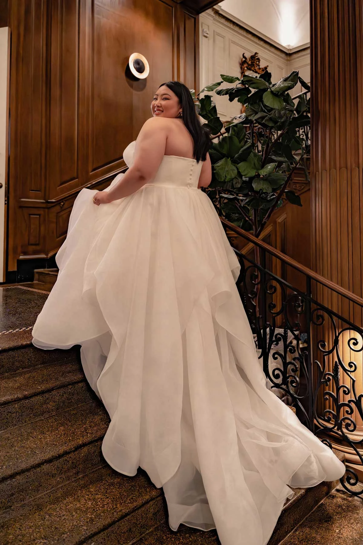 Bride wearing a plus size strapless ball gown wedding dress with a layered tulle skirt while walking up an elegant staircase