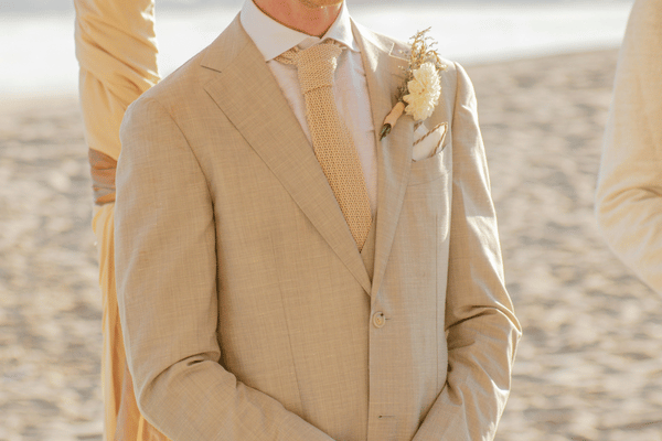 Beige-suited groom with boutonniere standing on a sunny beach, cropped at the face, with pale wedding attire in the background