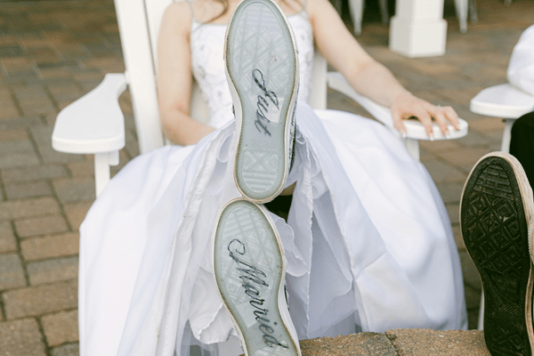 Bride in white dress lounges outdoors with feet up, showing Just Married on the soles of her shoes.