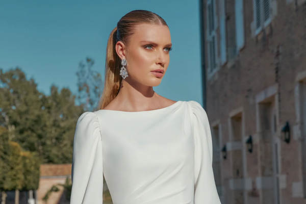 A woman in a white dress with puff sleeves, a Bateau (boat) neckline, styled hair, and earrings, posing outdoors.