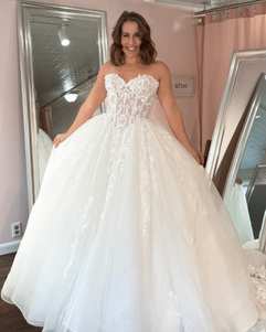Bride standing in a boutique wearing a strapless ball gown wedding dress with a structured bodice and full glitter tulle skirt, smiling with arms slightly out to showcase the volume.
