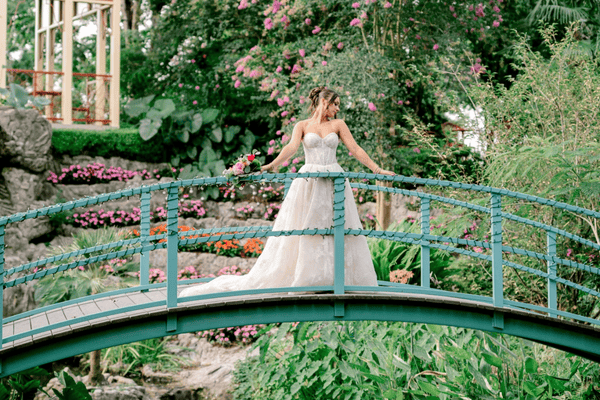 A bride in a white wedding dress stands on a blue bridge surrounded by colorful flowers and green plants at a wedding venue in Gulfport, MS.