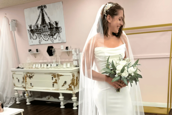 Smiling bride in white gown and veil holds bouquet in a pastel bridal boutique with chandelier art on the wall.