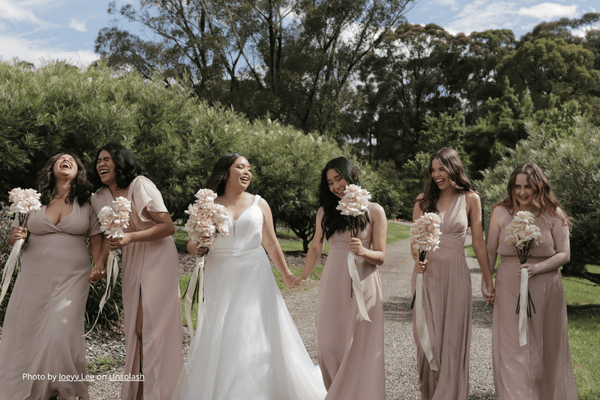 Six women in dresses walk joyfully on a garden path, holding bouquets. The bride is in white, bridesmaids in blush. Trees in the background.
