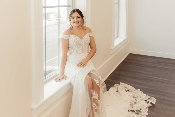 Bride in white dress sits by a large window, smiling. The gown has intricate lace patterns. Bright, airy room with wooden floor.