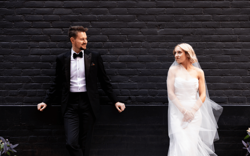 Bride and groom in wedding attire pose against a black brick wall, smiling at each other.
